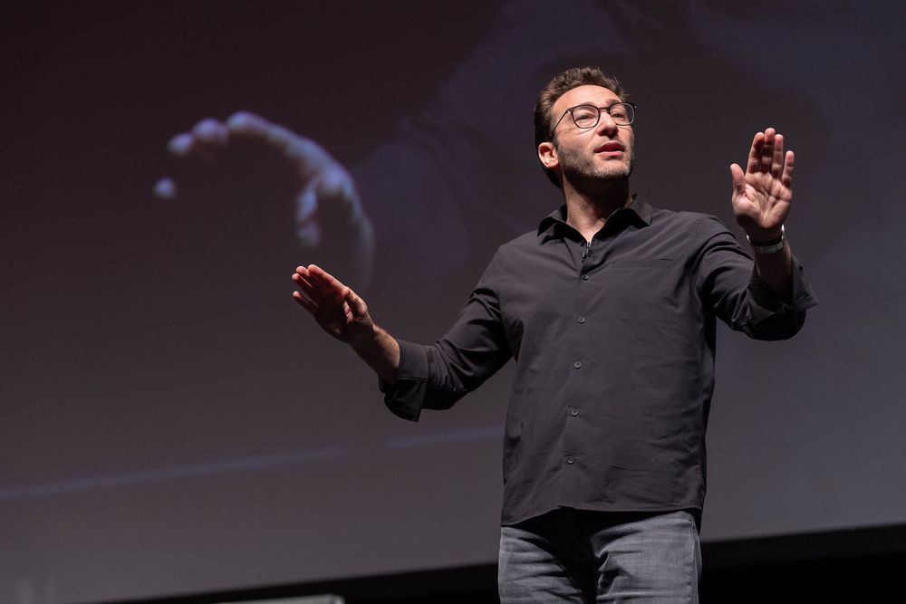 A presenter wearing a dark blazer gestures while speaking on stage at a technology conference.