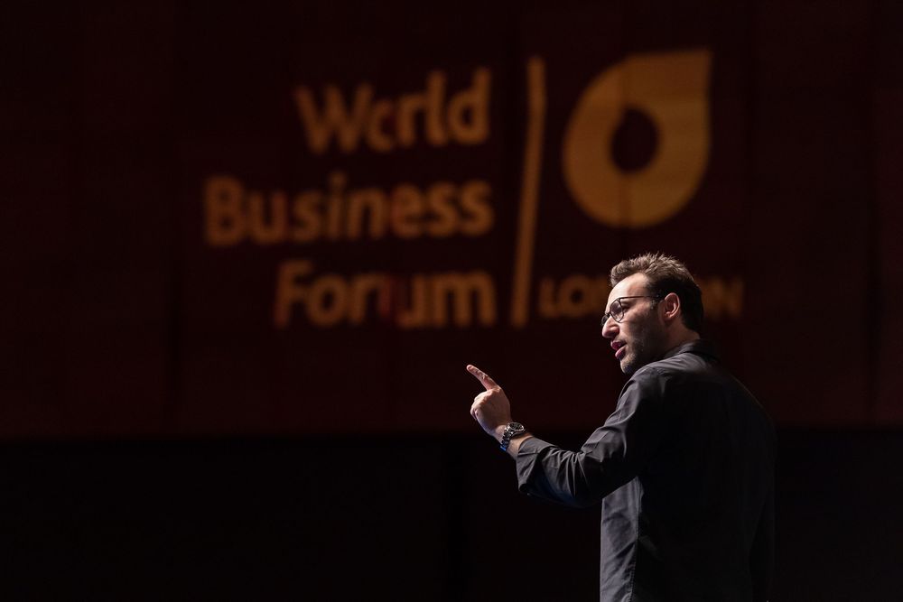 A person gestures while speaking at the Business Forum event with dark backdrop and illuminated signage.