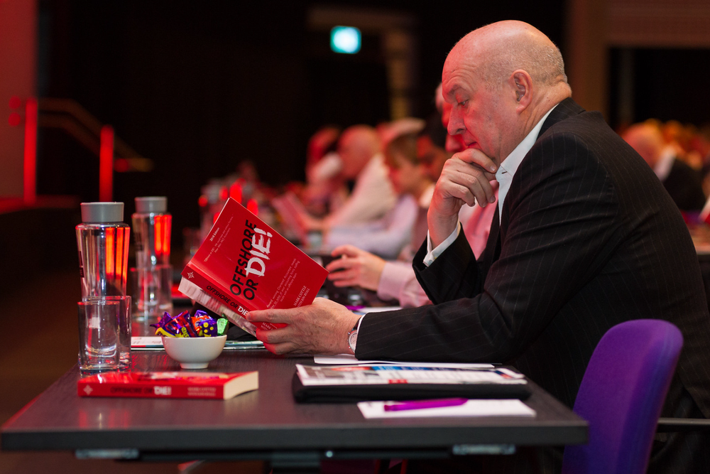 A person in formal attire sits at a desk with a red book and glass of water during an indoor event.