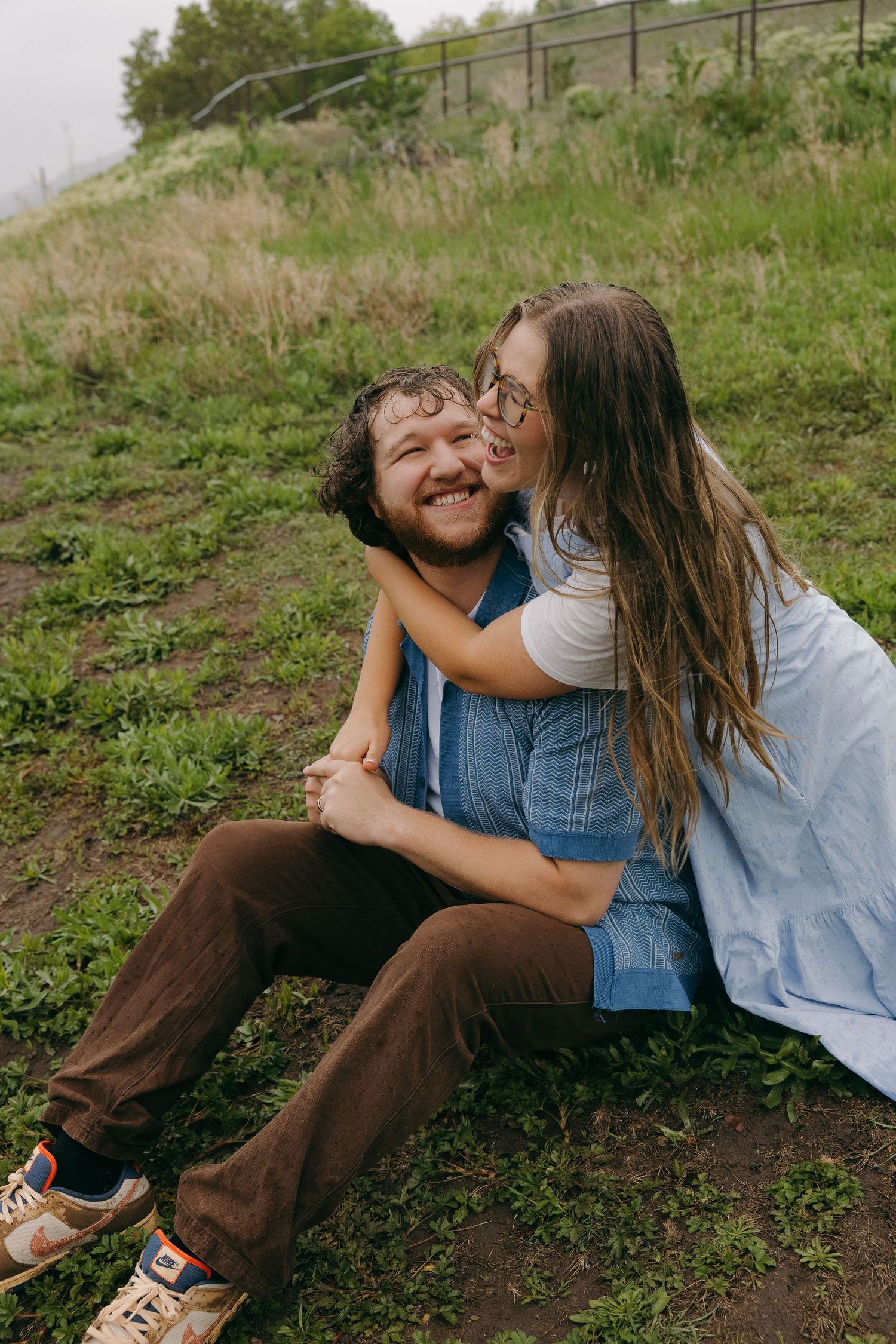 Couple sitting on grass, woman hugging man from behind and kissing his cheek outdoors.