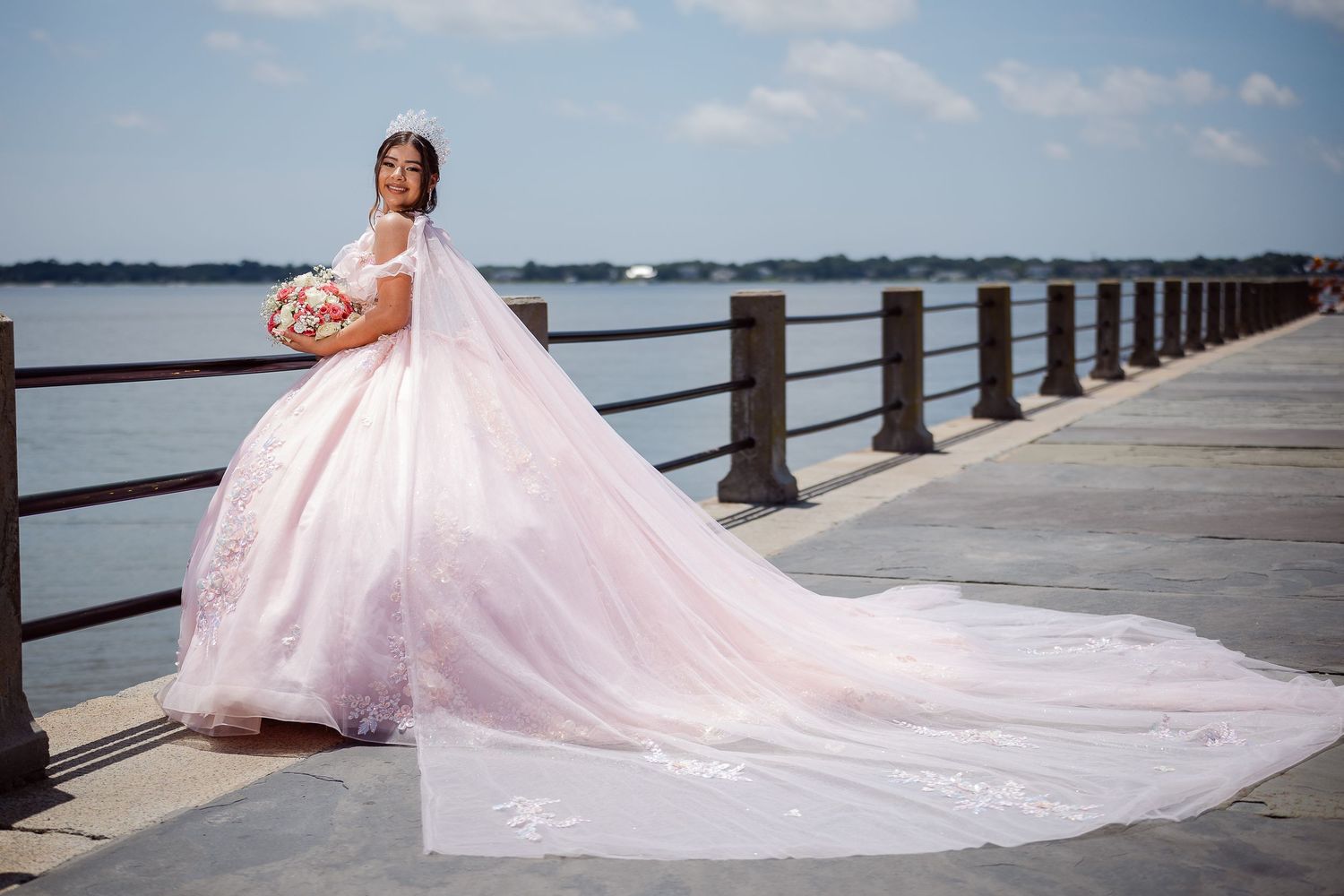 A  quinceañera in a light pink ball gown with a flowing train poses on a waterfront pier against a cloudy blue sky.