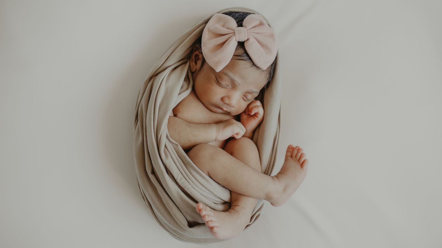 Peaceful newborn baby sleeping curled up in a beige wrap with a pink bow headband on a white background.