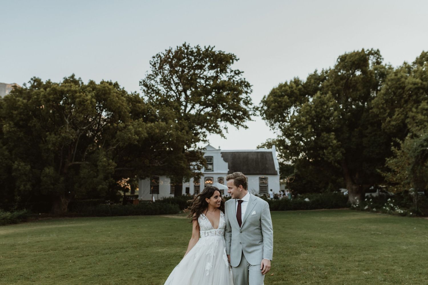 Newlyweds stand together on green lawn in front of white farmhouse.