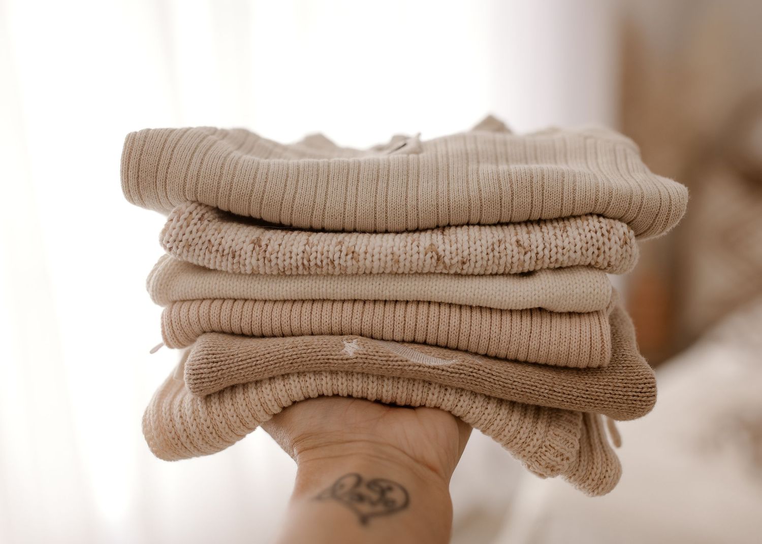 A neatly folded stack of beige knitted sweaters held by hands with a small tattoo visible.
