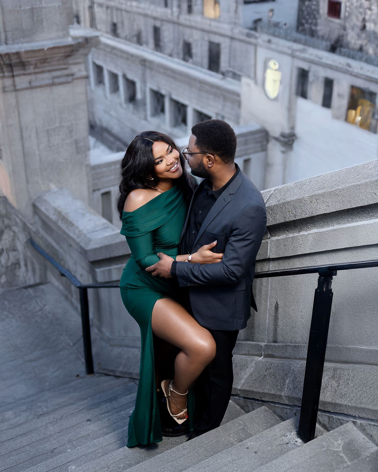 A romantic couple embraces on stone steps, with the woman wearing an elegant emerald green dress and the man in a dark blazer.