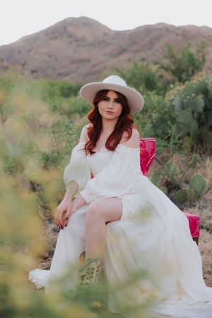Flowing white dress and wide brim hat in desert maternity photoshoot with pink vintage chair.
