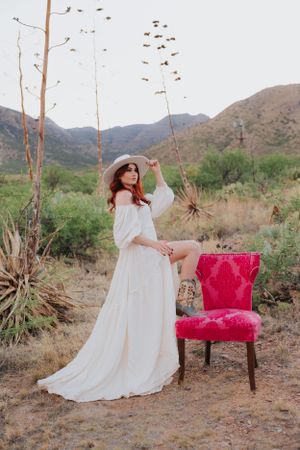 White flowy dress contrasts with bright pink vintage chair against desert mountain backdrop.