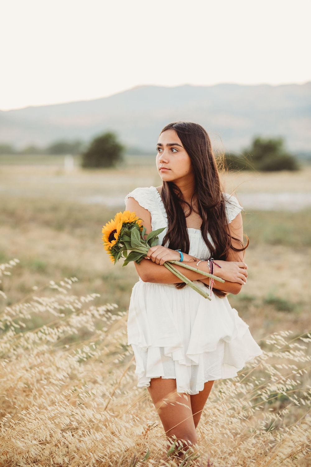 A person in a white summer dress holding sunflowers stands in a golden field during golden hour.