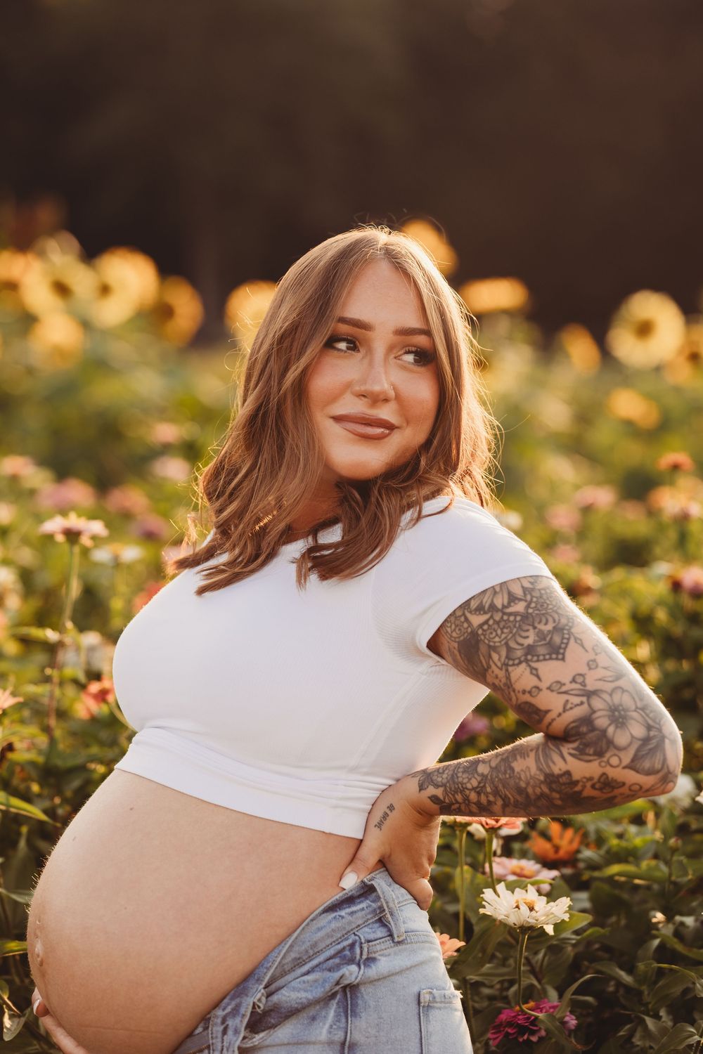 A pregnant woman in a white crop top stands in a sunlit flower garden with visible arm tattoos and denim shorts.
