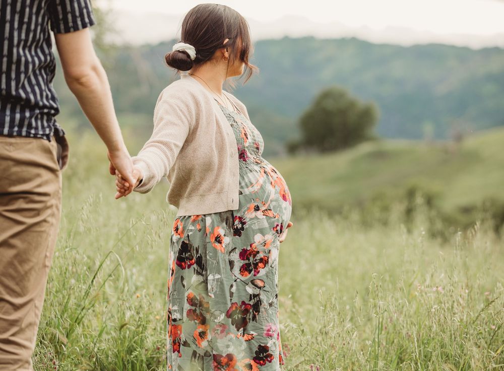 Couple holds hands while walking together through a sunny meadow with tall grass and rolling hills in background.
