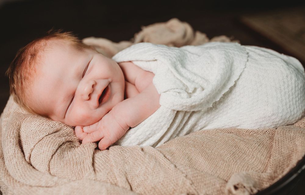 A peaceful baby sleeping while wrapped in white swaddle blankets on a soft beige textured surface.