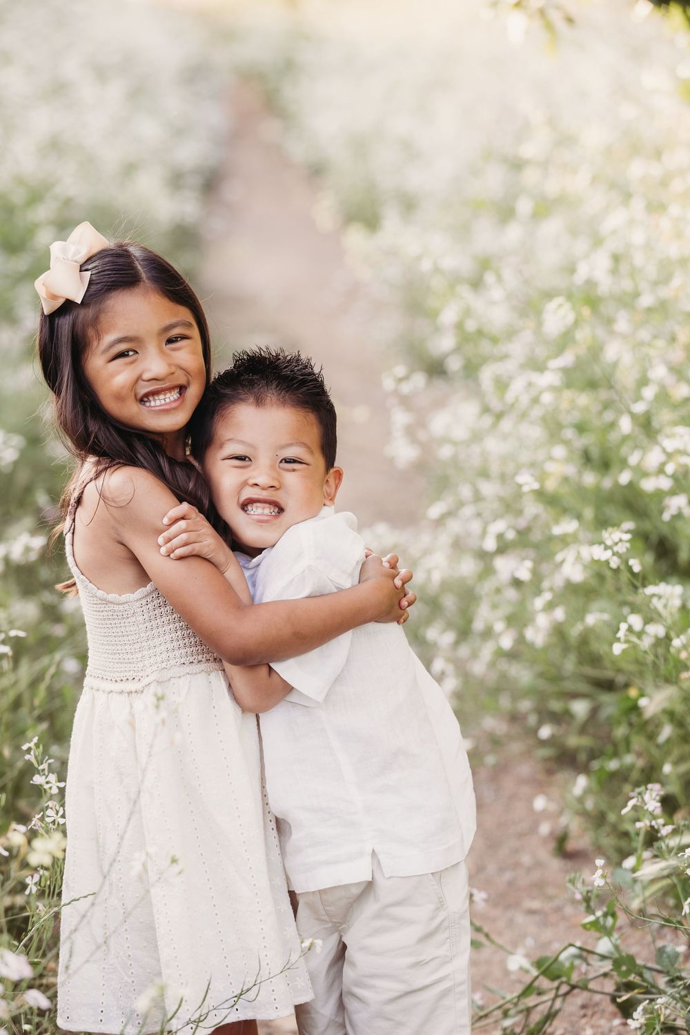 Siblings in white outfits play and smile together among blooming white flowers in an outdoor garden setting.