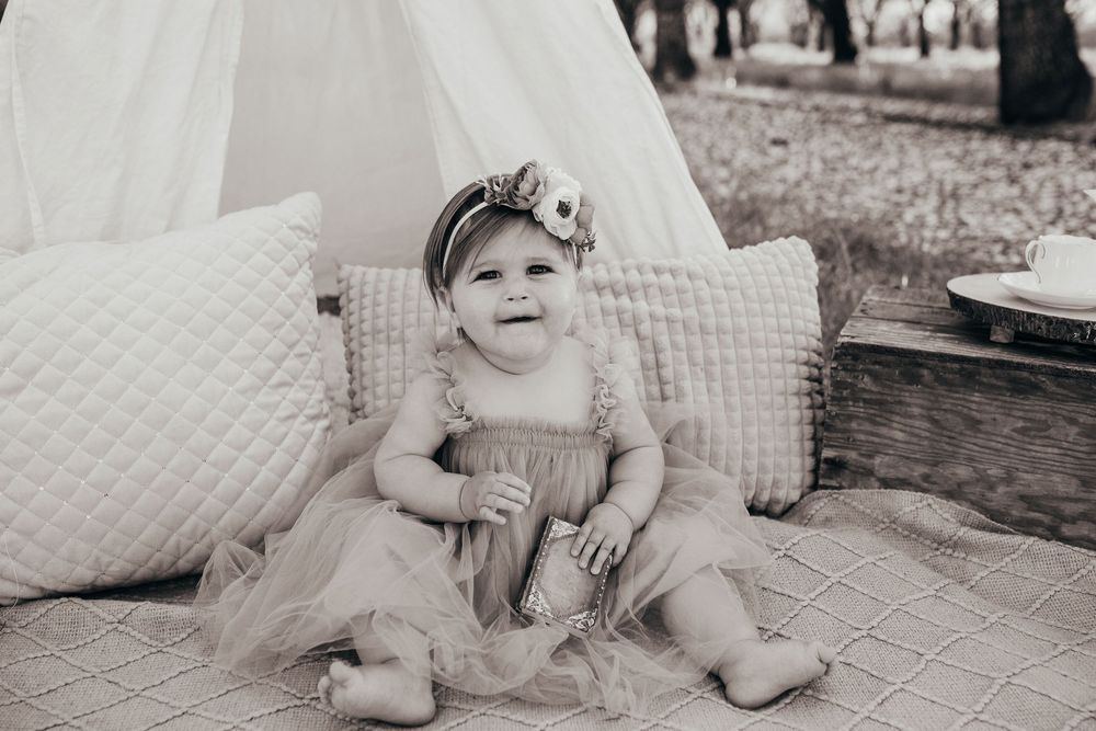 Black and white photo of small baby wearing dress sitting on cushions near teepee outdoors.