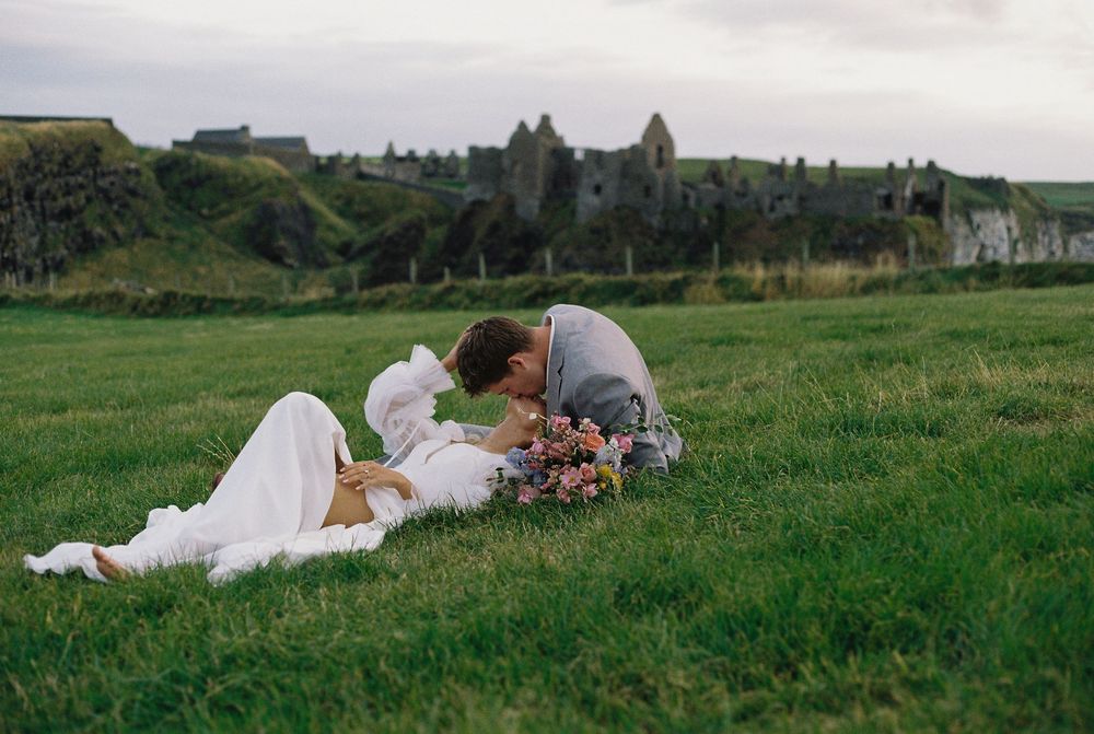 Couple eloping at Dunluce Castle in Northern Ireland laying on the grass with the castle behind them