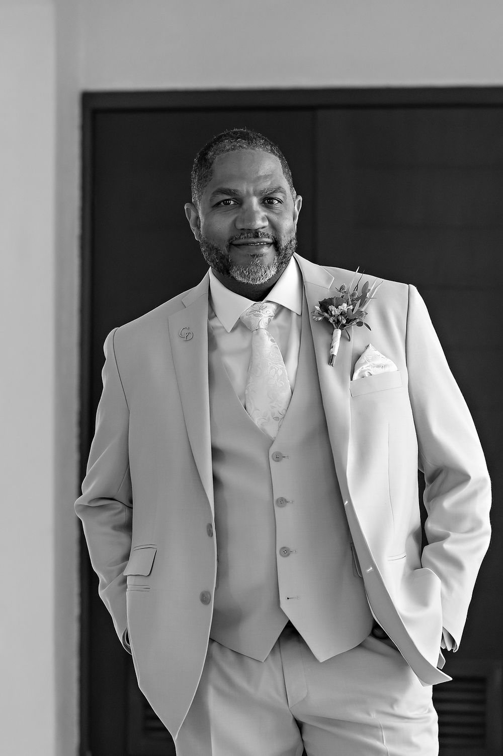 A person wearing a light-colored three-piece suit with a boutonniere poses with hands in pockets in black and white.