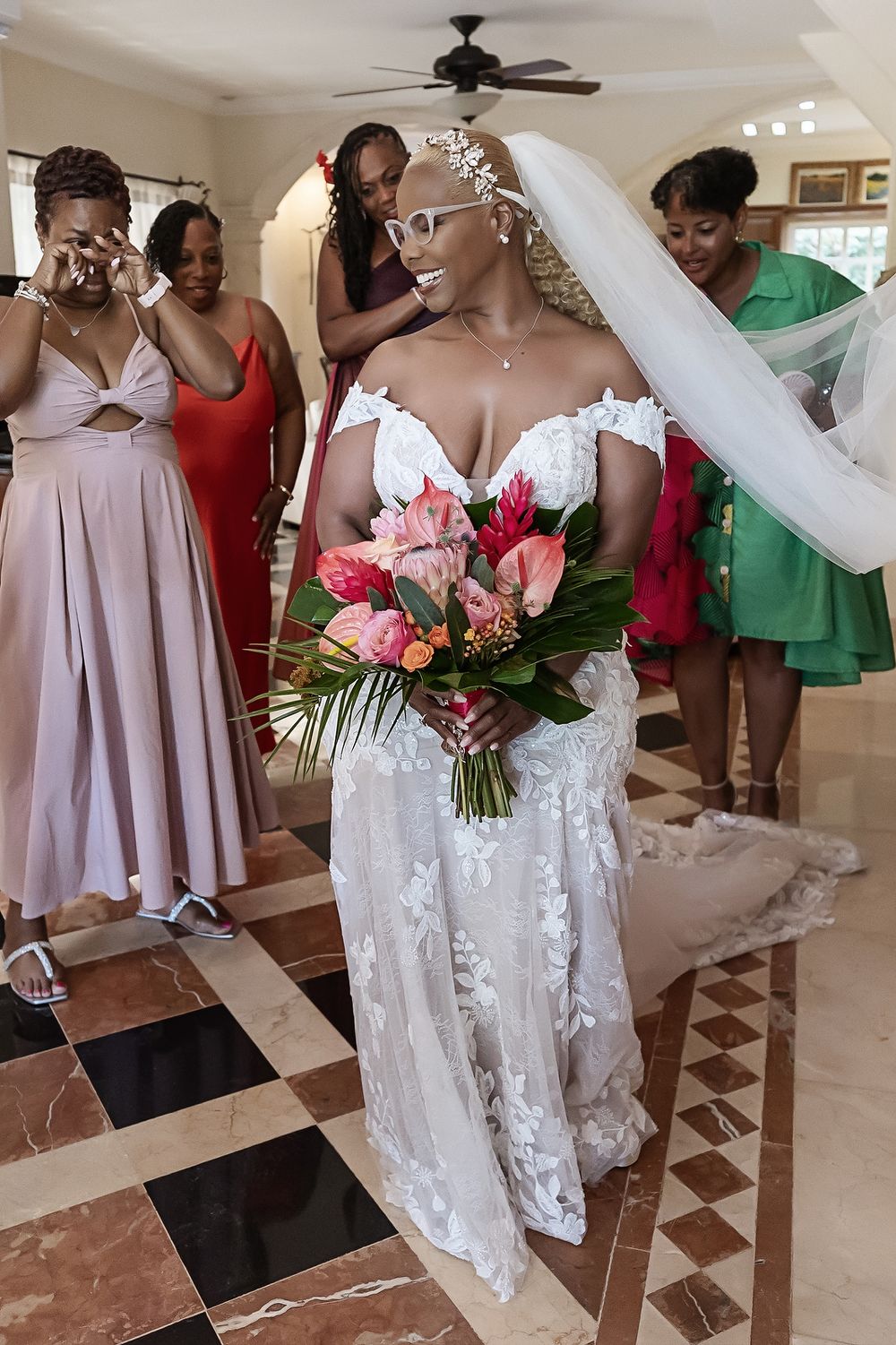 Bridal party members in lilac dresses and a bride in white lace gown gather together holding vibrant floral bouquets.