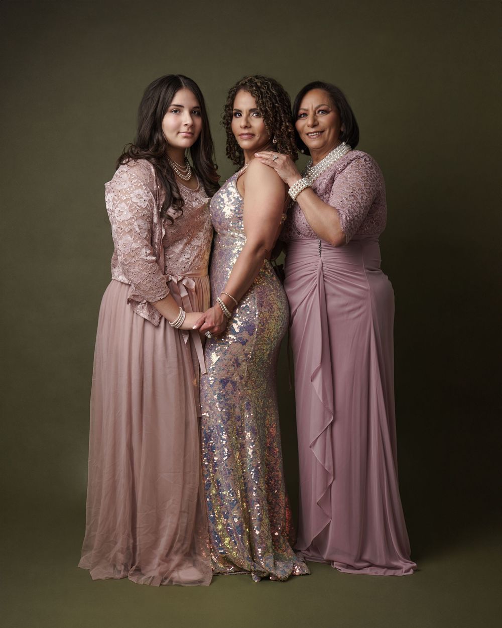Three people in elegant evening gowns pose together against dark backdrop in glamorous portrait session.