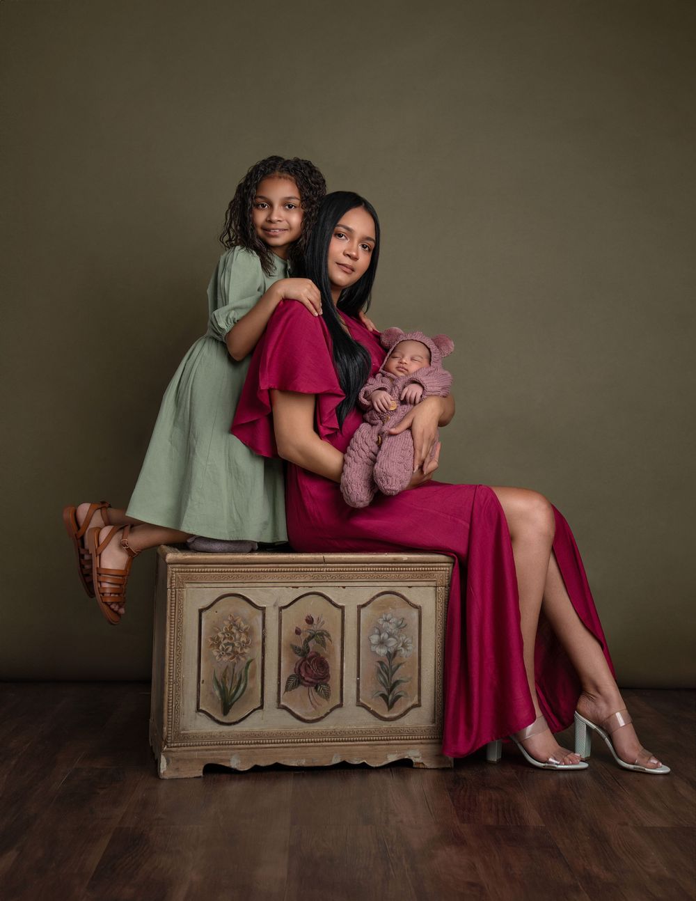 Portrait of figures in elegant dresses posing together on an antique wooden chest against olive backdrop.