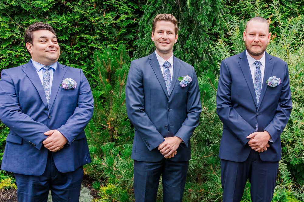 A lineup of groomsmen in navy blue suits stand against a lush green garden backdrop at an outdoor wedding venue.