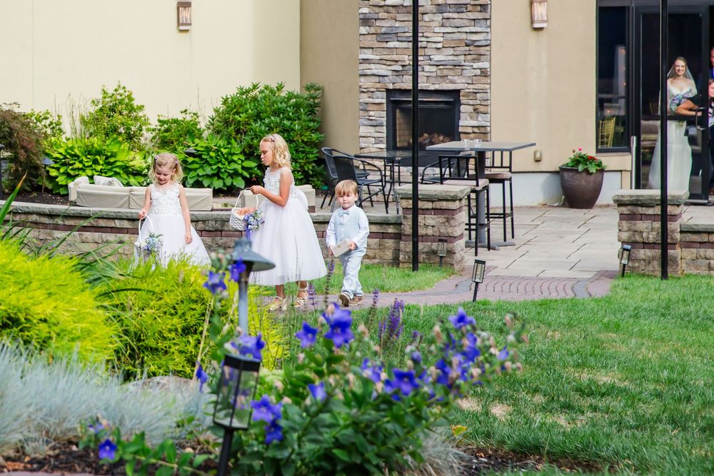 Children run and play on a patio area near purple flowers and green landscaping on a sunny day.