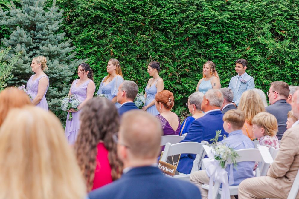 A wedding ceremony in an outdoor garden setting with guests seated on chairs and purple-clad attendants standing up front.