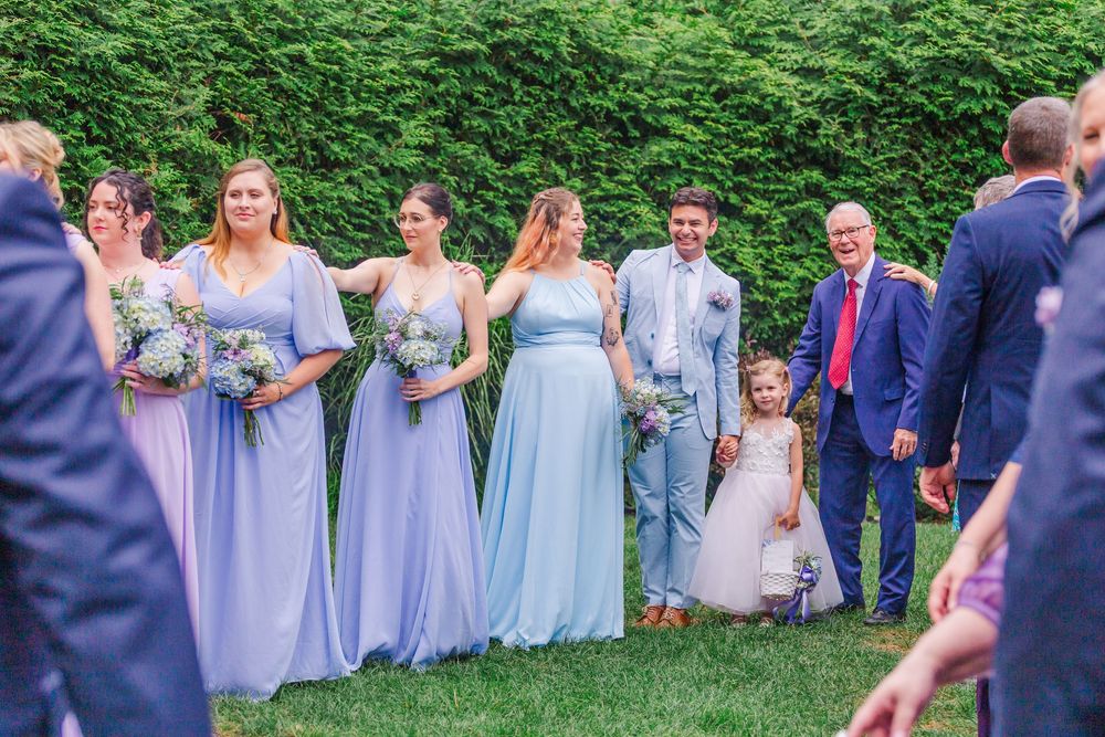 A wedding party in soft lilac and blue dresses poses together on green grass during an outdoor ceremony.