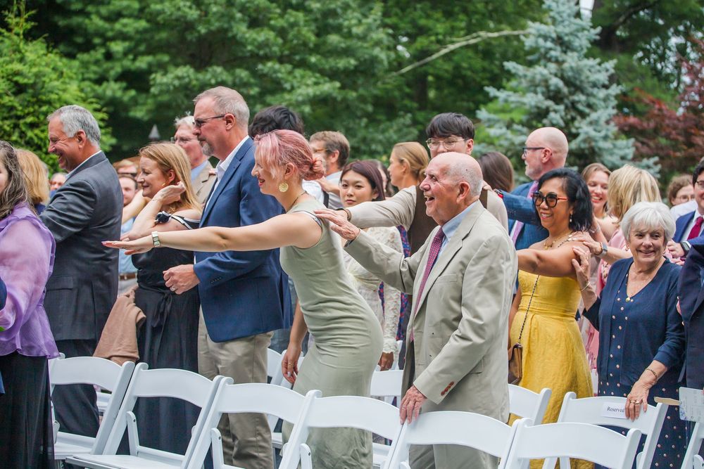 Wedding guests dance in celebration on an outdoor dance floor during a summer reception among trees.