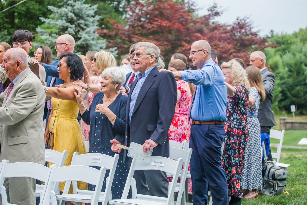 Wedding guests stand from their chairs and look into the distance together during an outdoor garden ceremony.