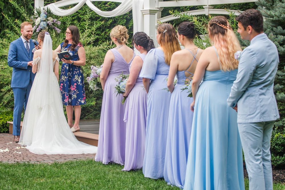 Wedding party stands in a garden setting with bridesmaids wearing coordinated purple and blue floor-length gowns.
