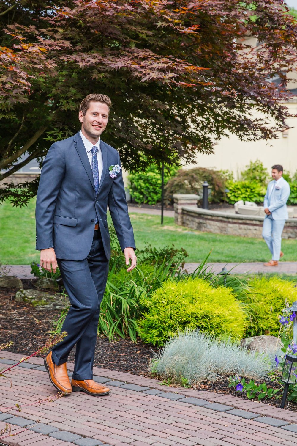A well-dressed person in a navy suit and tan shoes stands on a brick path in a landscaped garden with Japanese maple trees.