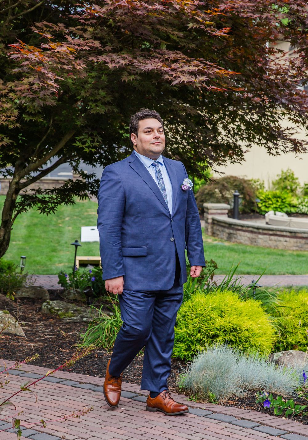 Groom in navy blue suit and brown shoes poses for wedding photos on brick pathway in garden setting.