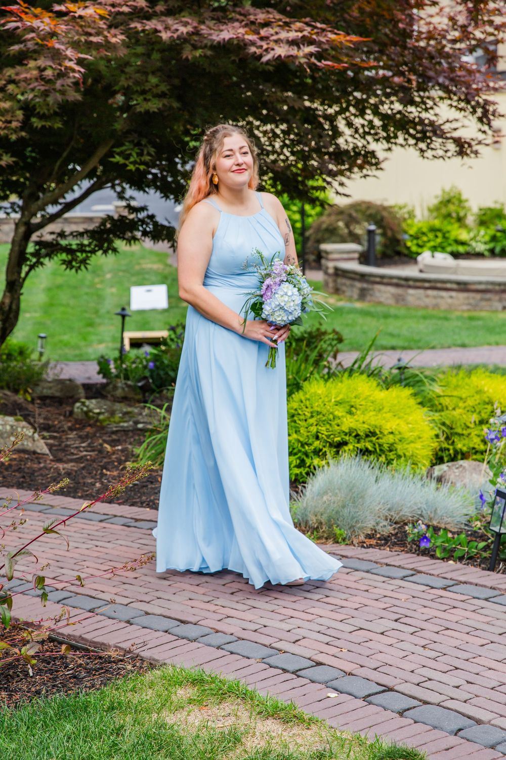 Someone in a flowing light blue bridesmaid dress holds a bouquet while standing on a brick path in a garden setting.