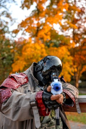 An airsoft or paintball player in protective gear aims a blue-tipped gun against a backdrop of vibrant autumn foliage.