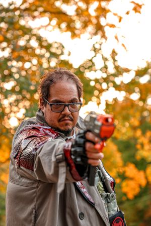 Someone aims a red toy Nerf gun while wearing glasses and a coat against orange autumn foliage background.