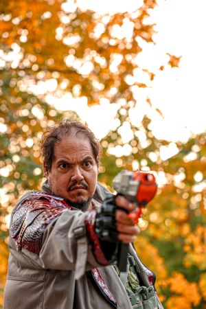 A person in autumn gear aims a red paintball gun against a backdrop of orange fall foliage at sunset.