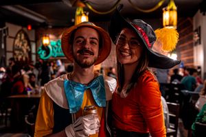Two people in colorful Halloween costumes smile together at a bar with warm lighting and festive decorations.