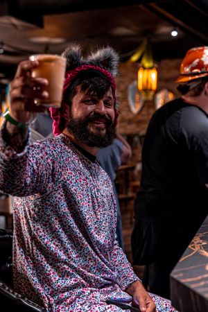 Person wearing a floral print outfit and mouse ears raises a drink with a smile at what appears to be a bar setting.