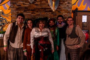 A group of people pose together in medieval and renaissance costumes against a brick wall with orange decorative flames.