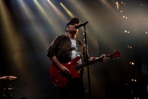 A musician performs on stage with a red electric guitar under dramatic spotlights during a concert performance.