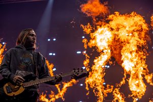 Musician plays electric bass on stage with dramatic flames erupting in the background during a live performance.