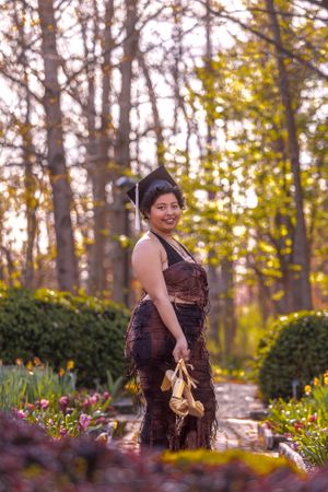 Graduate stands in a garden wearing brown dress holding graduation shoes at sunset among blooming flowers and trees.
