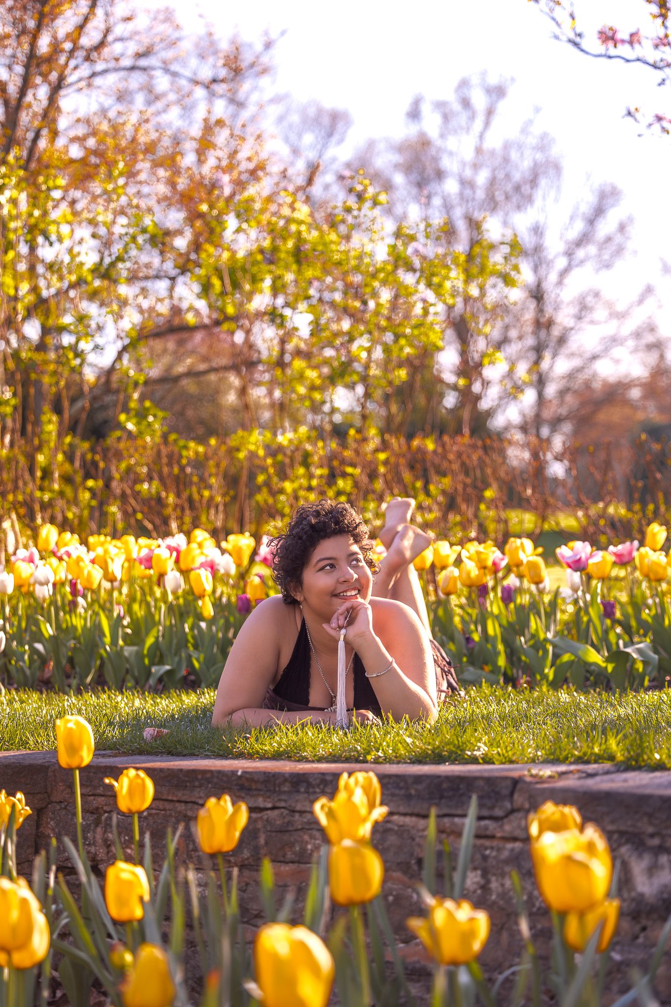 A person poses laying in a garden surrounded by bright yellow tulips on a sunny spring day.