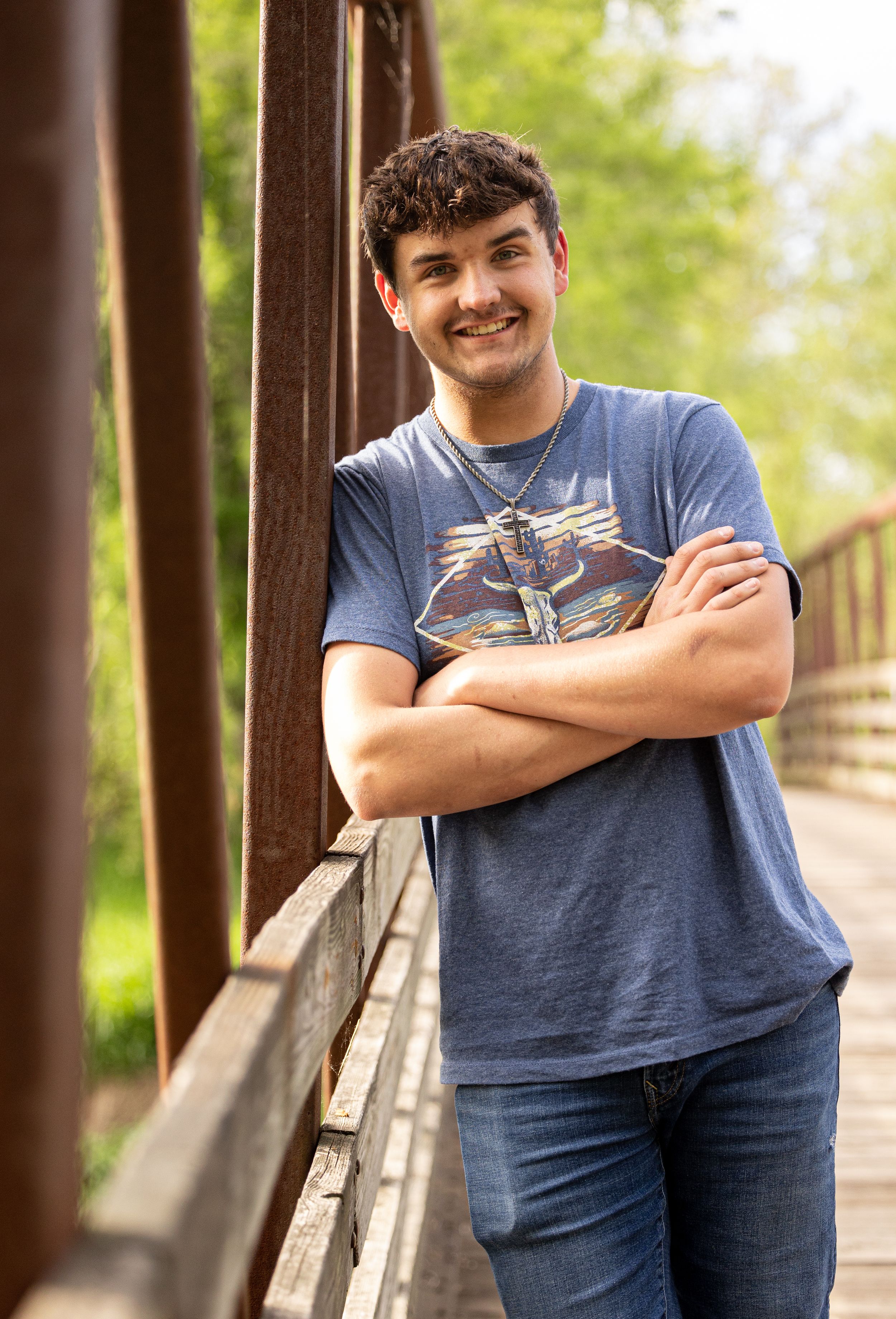 Young person in a blue graphic t-shirt smiling while standing on a wooden bridge with a blurred natural background.