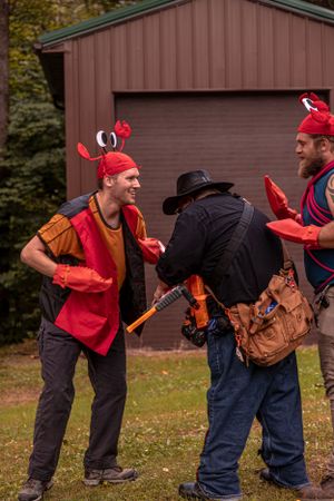 A small group wearing red vests and hats gather outdoors near a brown barn while holding tools and gear.