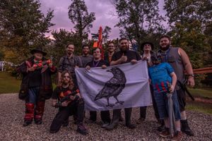 A group of people pose outdoors at dusk holding a flag with a bird design against a purple-hued sky and trees.