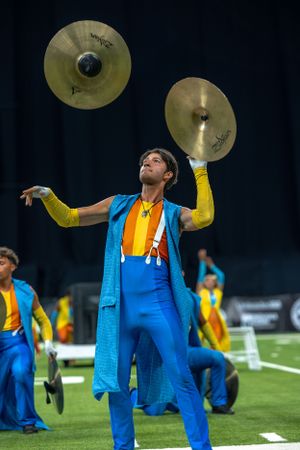 Performer in bright blue jumpsuit and yellow sleeves holding cymbals during marching band performance on field.