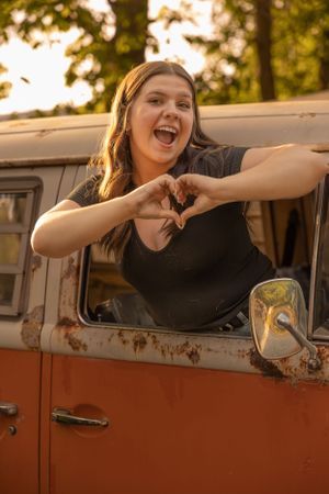 Someone leans out of a rusty orange vintage VW van window making a heart shape with their hands during golden hour.