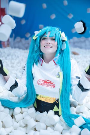 Cosplayer with bright turquoise hair wearing a white jacket poses playfully in a pile of marshmallows against a blue backdrop.