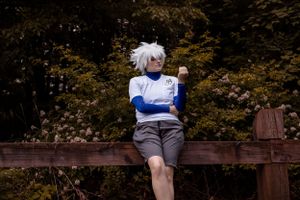 Anime cosplay character sitting on wooden railing in front of autumn foliage at dusk.
