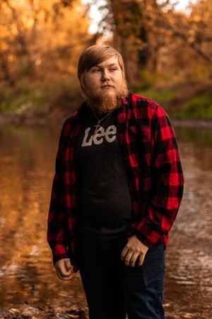 Person in red buffalo plaid flannel and Lee t-shirt poses near water during golden hour in autumn forest.